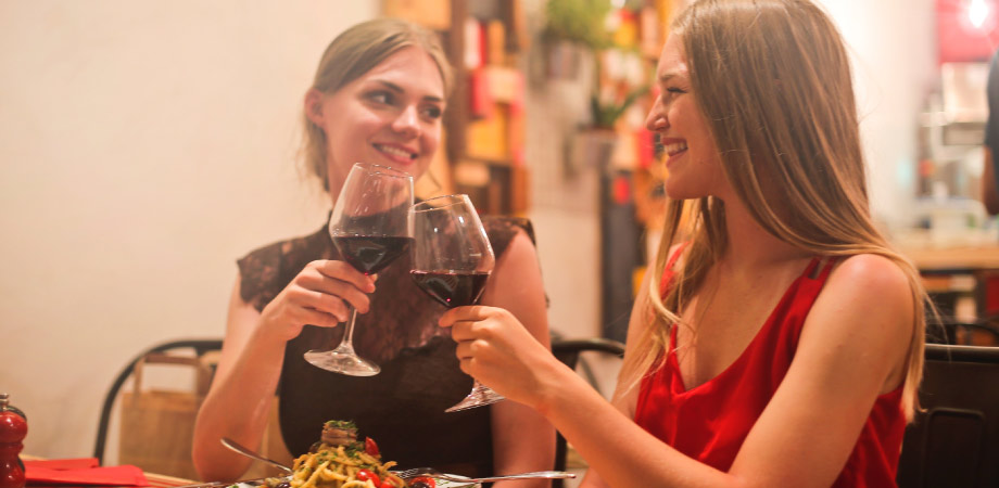 two women smiling at each other toasting with glasses of red wine